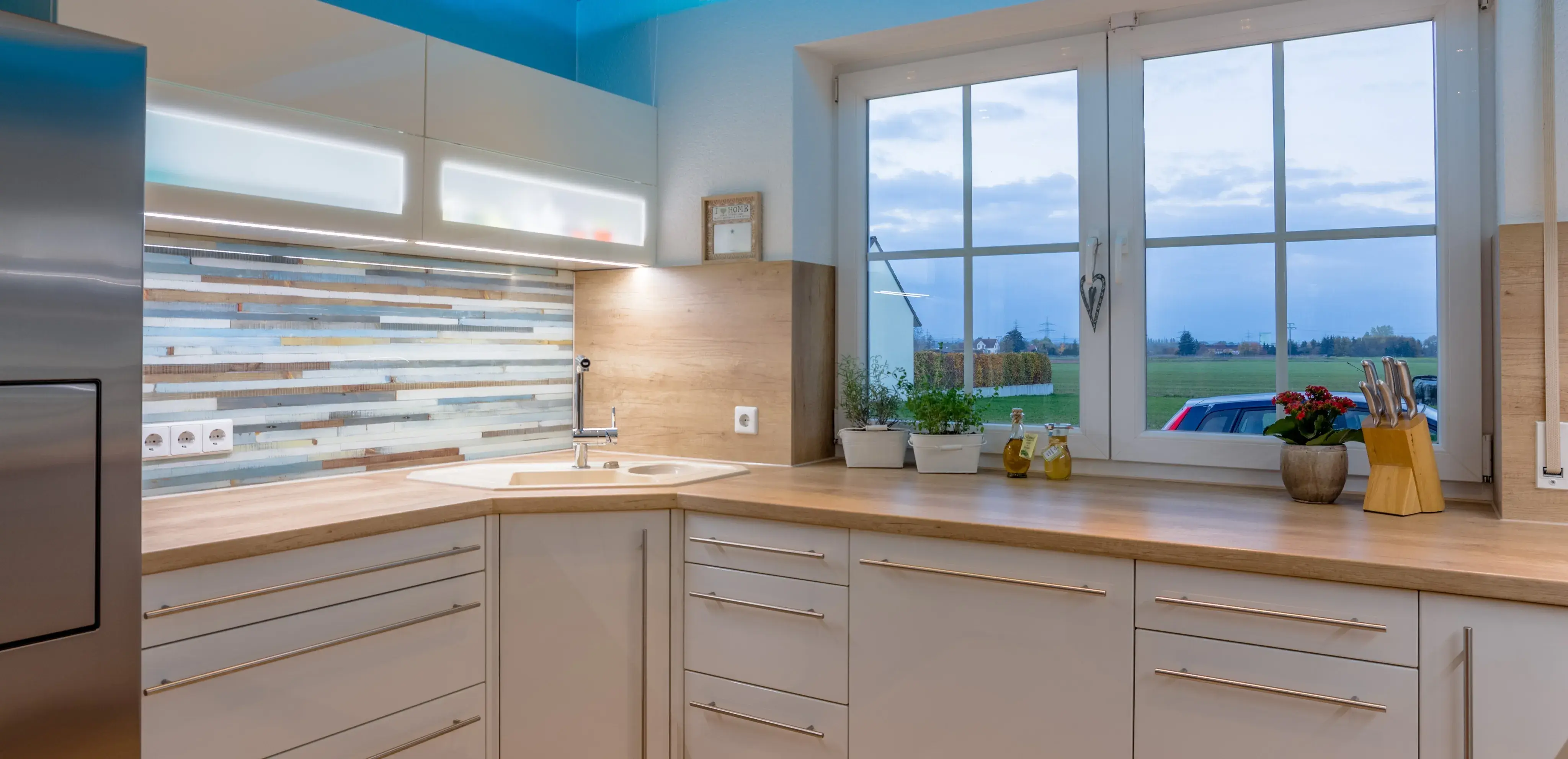 Moderne Küche mit Holzarbeitsplatten, großen Fenstern und Blick auf Felder bei Sonnenuntergang.