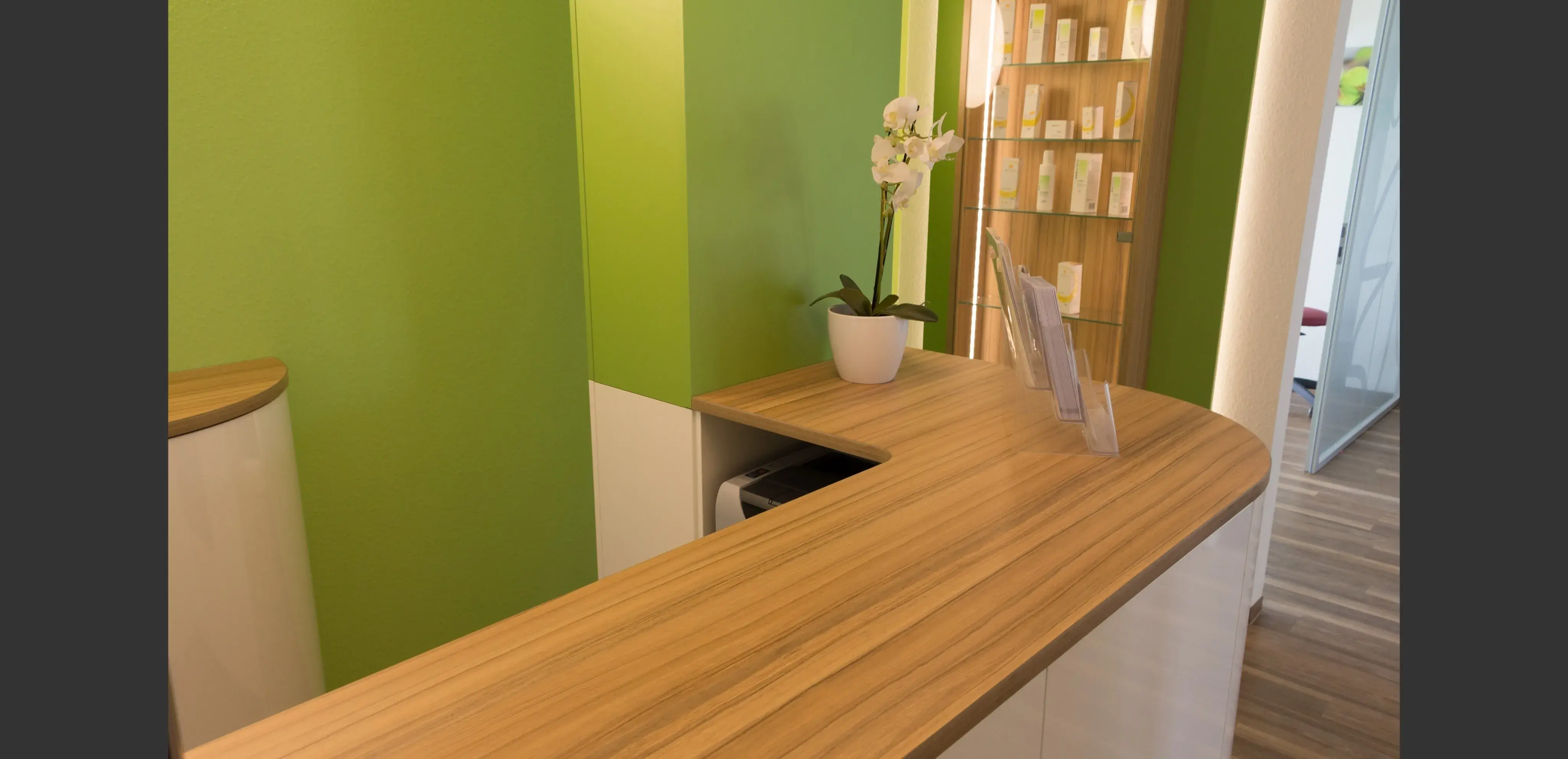 Heller Empfangstresen mit Holzoberfläche, weißer Orchidee und grüner Wand im Hintergrund.