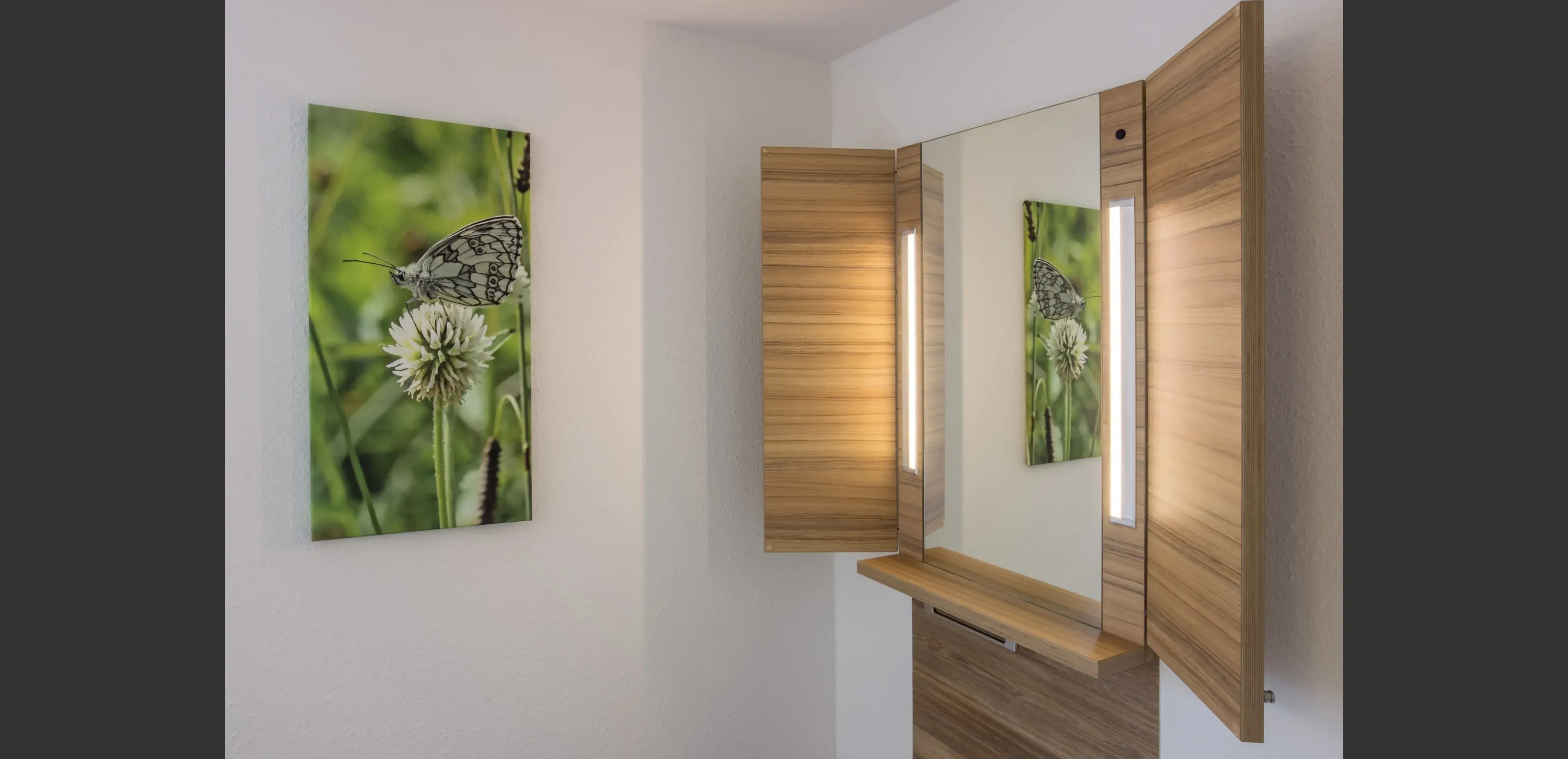 Spiegel mit Holzrahmen, daneben Leinwand mit Schmetterling auf Blume an weißer Wand.