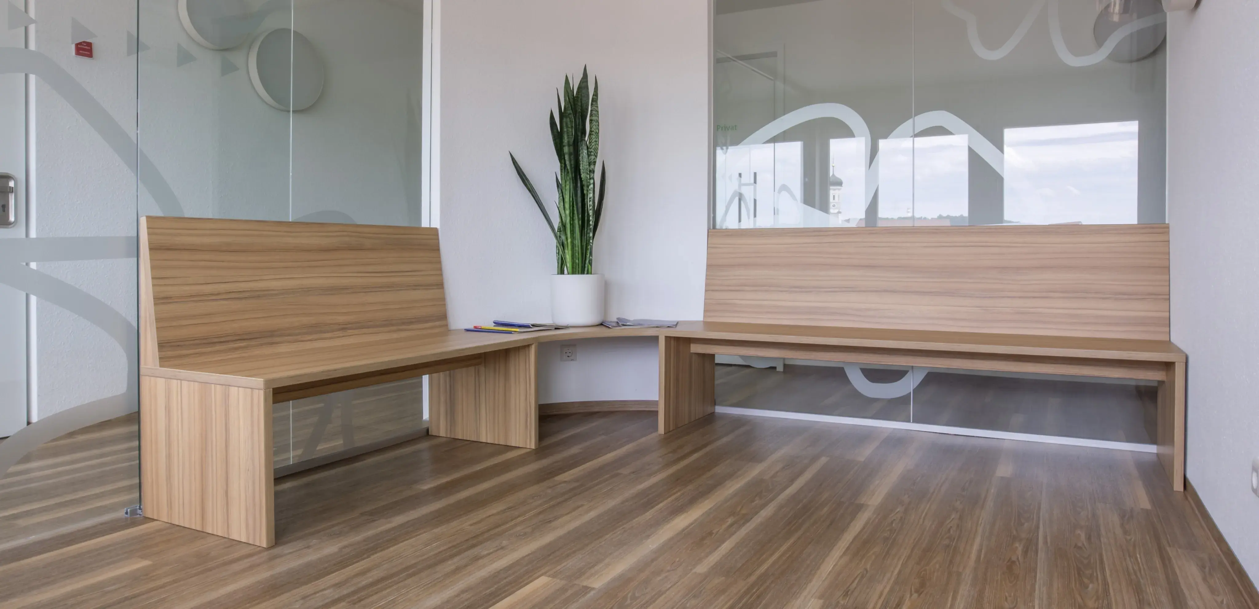 Warteraum mit Holzbänken, Pflanze in weißem Topf und Glaswänden.