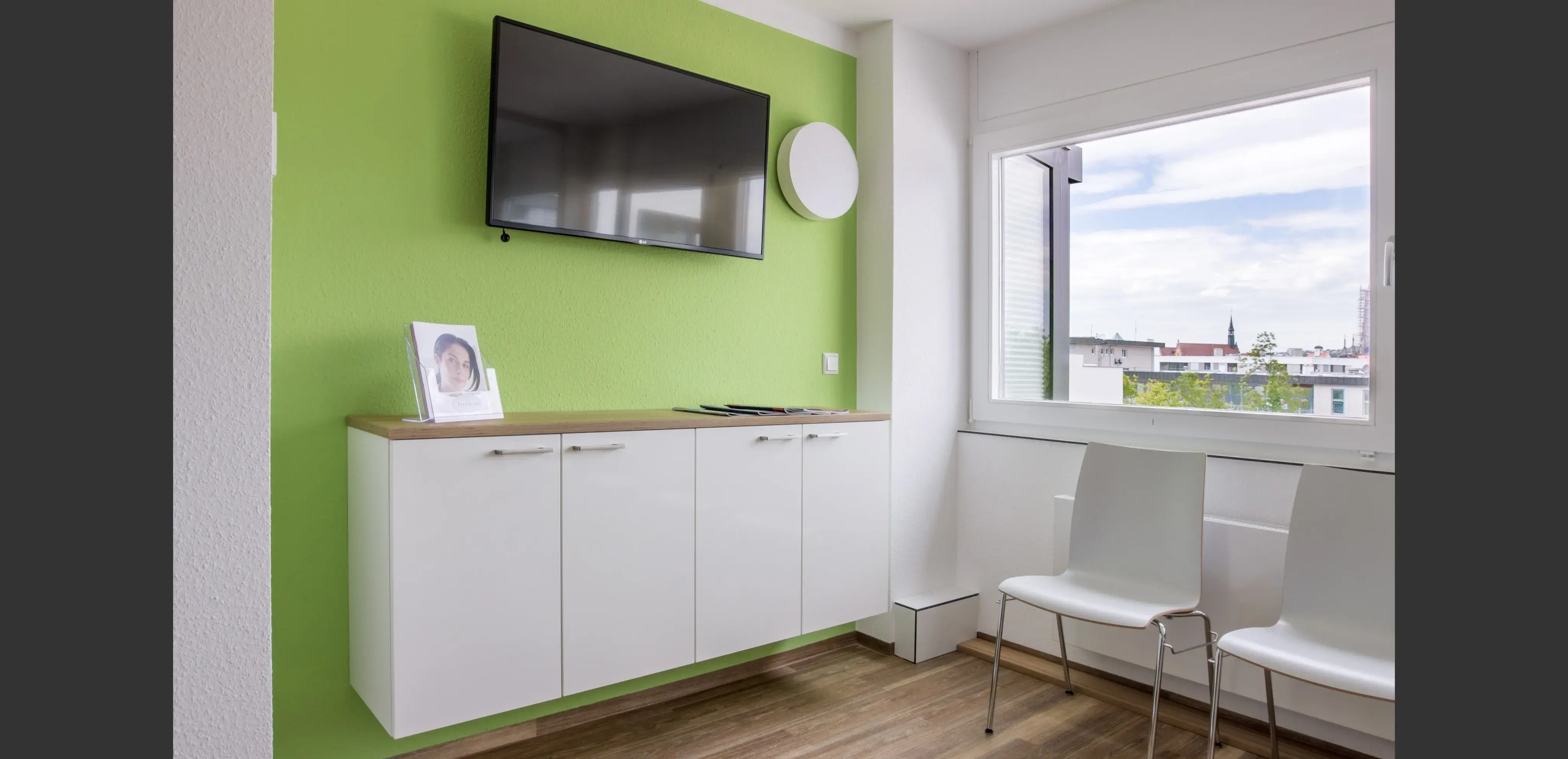 Heller Warteraum mit grüner Wand, weißen Stühlen, Fenster und Fernseher.