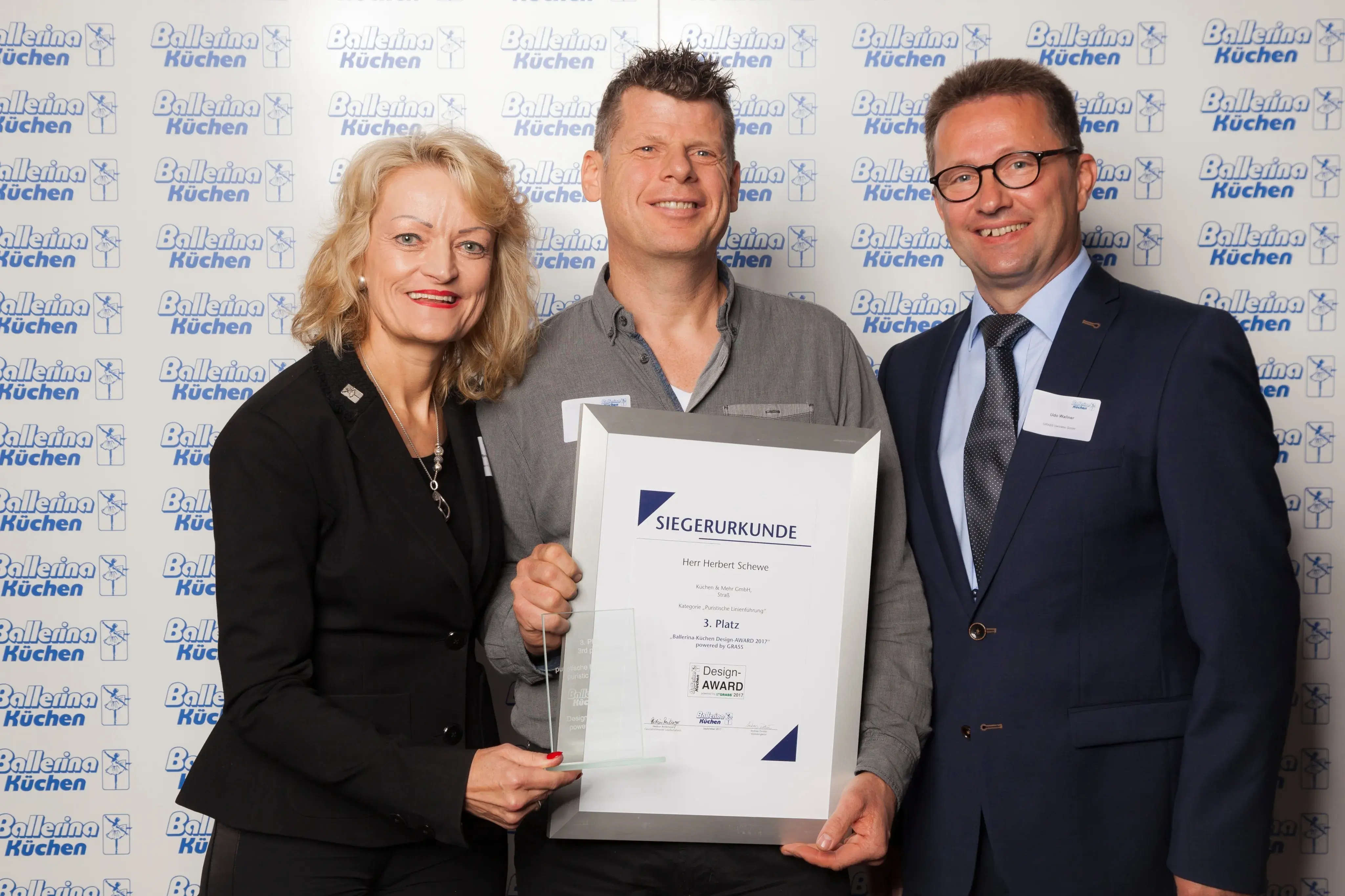 Drei Personen in Businesskleidung halten eine Urkunde und lächeln in die Kamera.