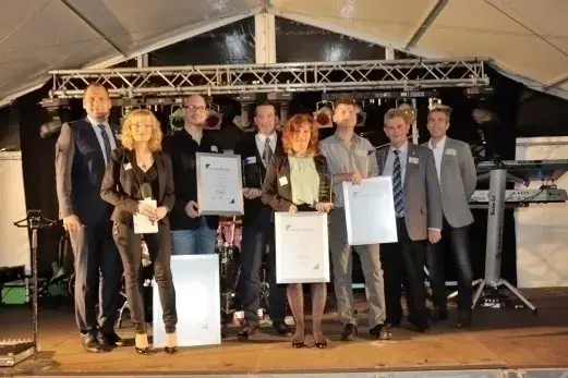 Gruppe von acht Personen steht auf einer Bühne und hält Urkunden in den Händen.