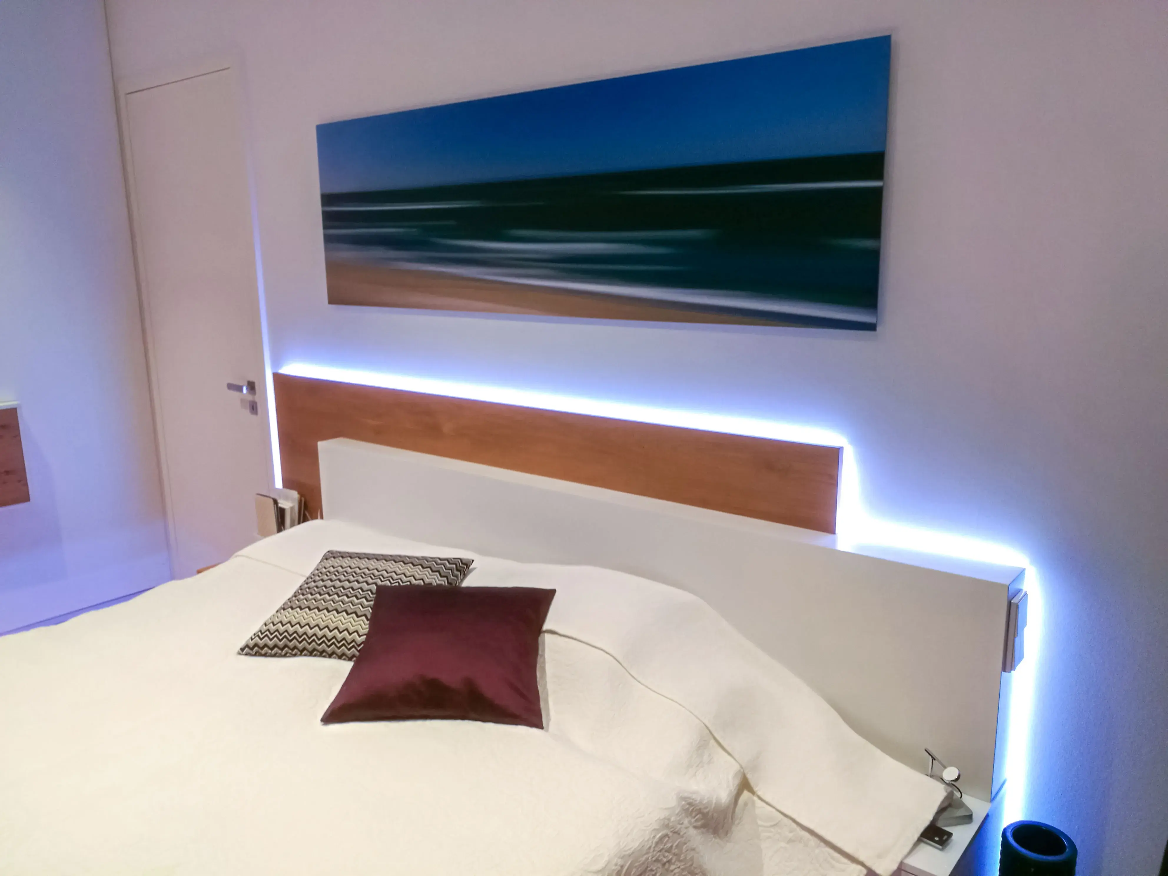 Modernes Schlafzimmer mit beleuchtetem Bettkopfteil und abstraktem Meeresbild an der Wand.