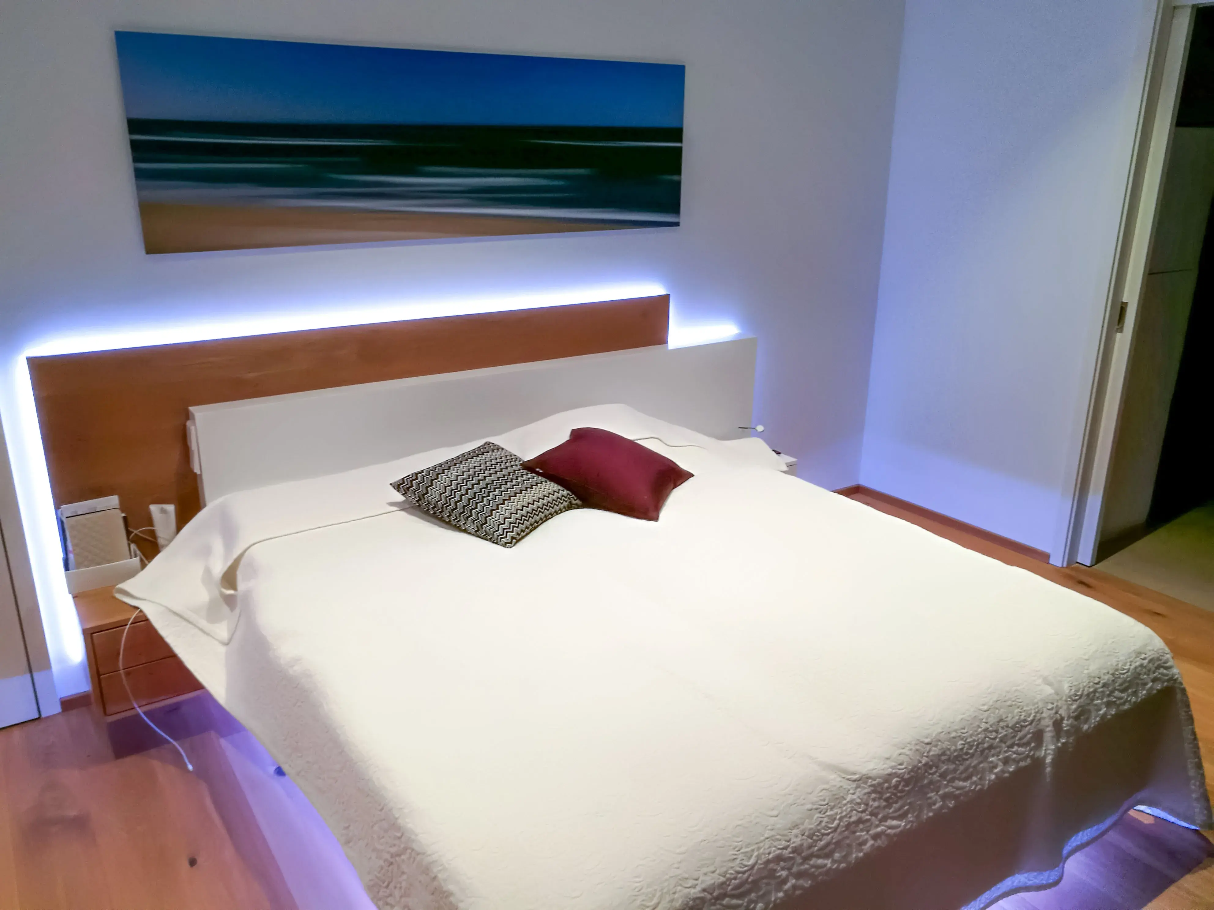 Modernes Schlafzimmer mit großem Bett, weißen Bettdecken und bunten Kissen, sanfte Beleuchtung.