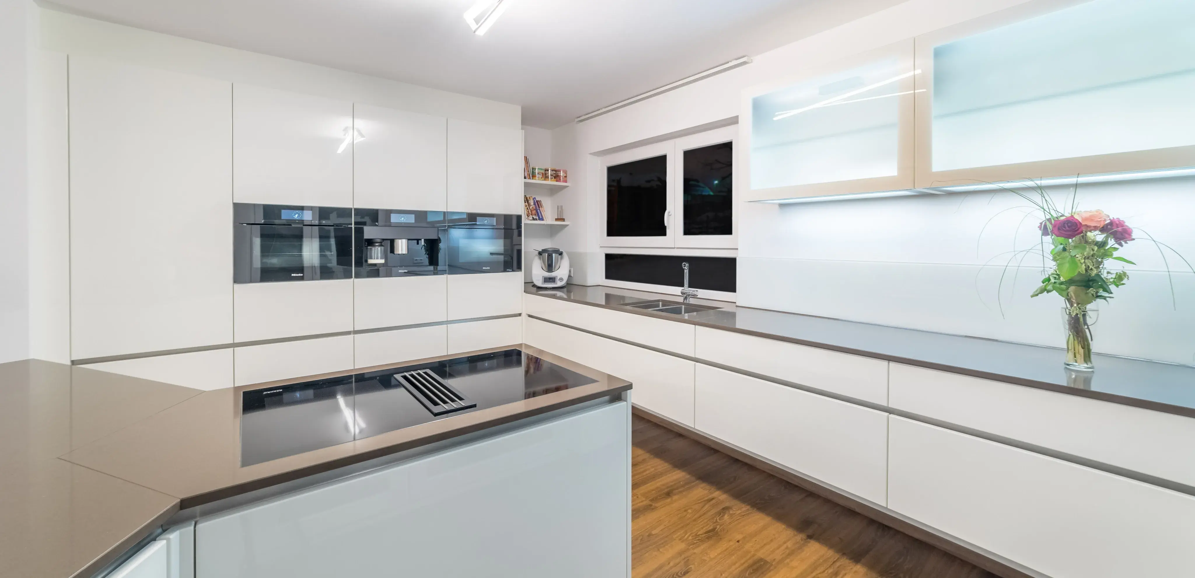 Moderne weiße Küche mit Kochinsel, Holzfußboden und Vase mit Blumen auf der Theke
