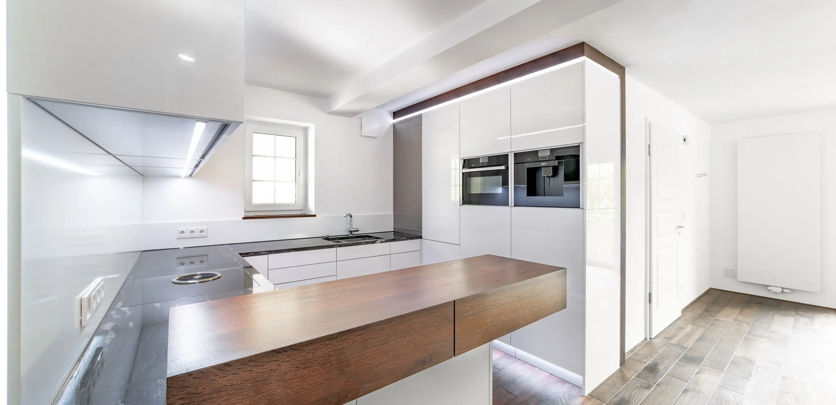 Moderne weiße Küche mit Holztheke und eingebauten Geräten, viel Tageslicht.