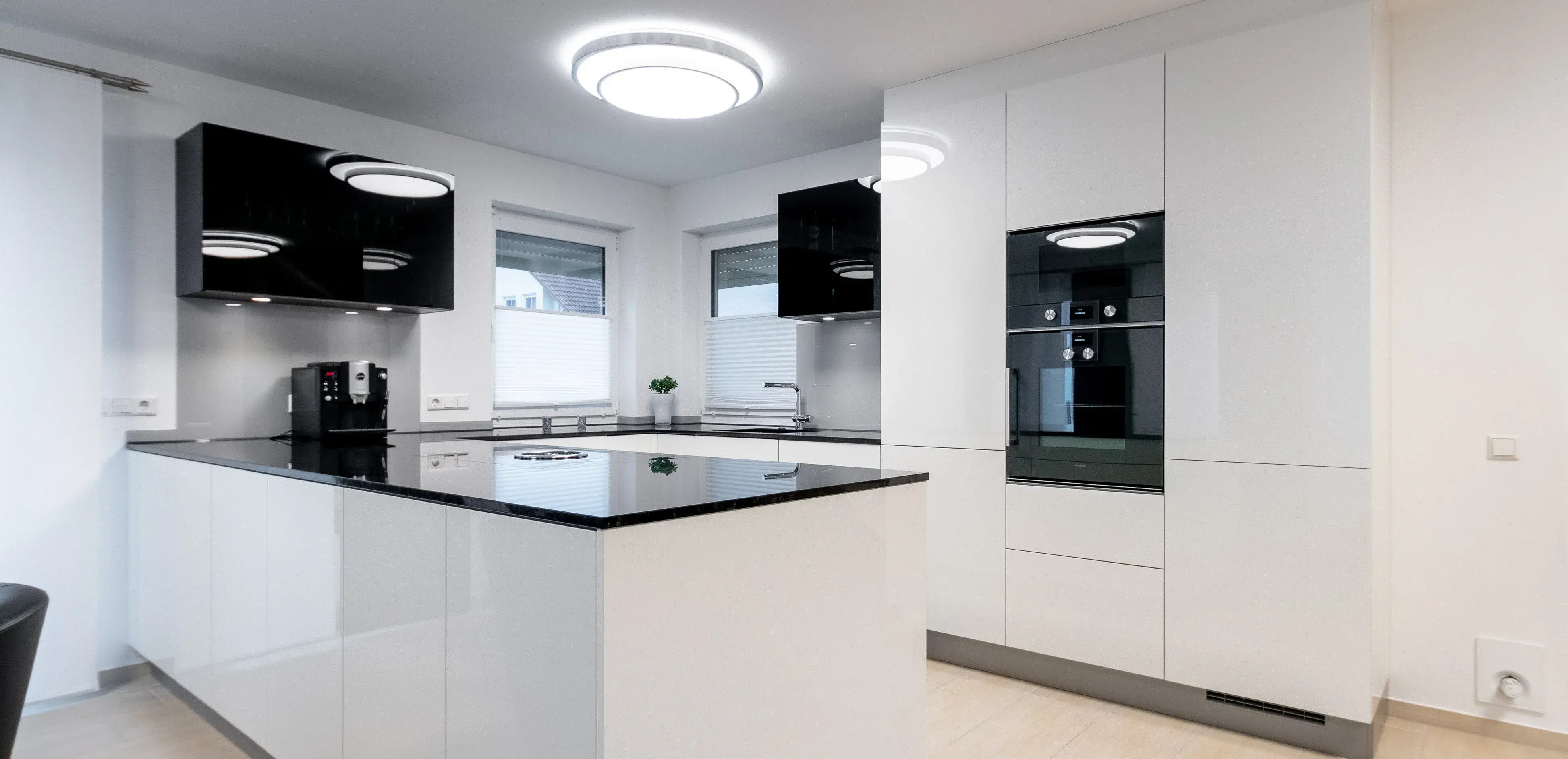 Moderne weiße Küche mit Hochglanzfronten, schwarzer Arbeitsplatte und hellem Boden.
