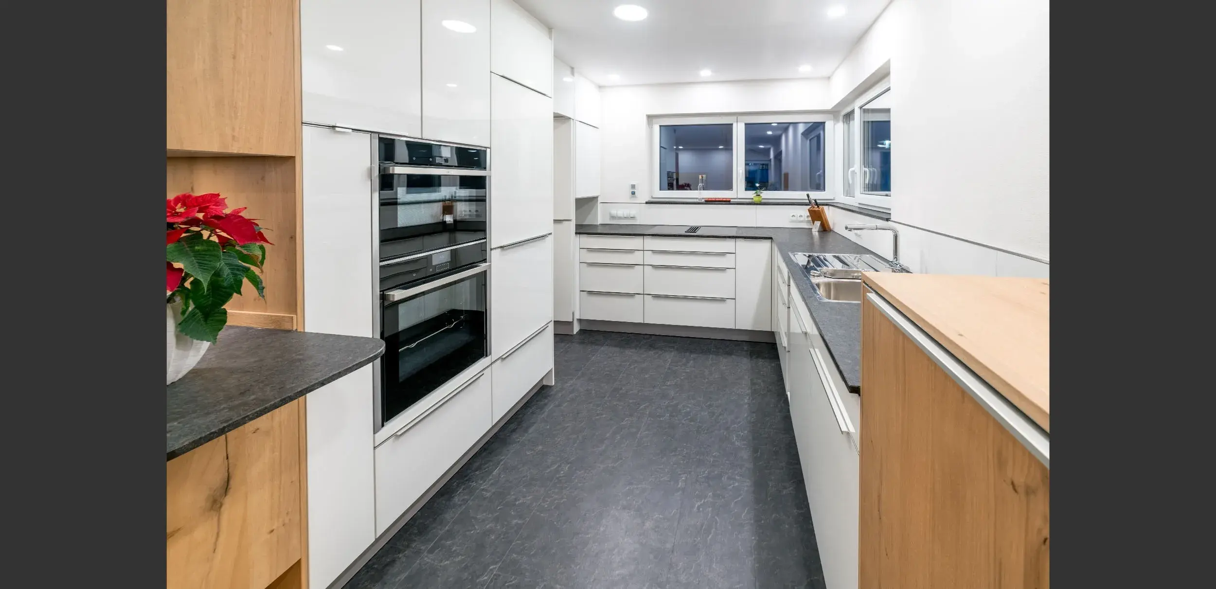 Moderne weiße Küche mit dunklem Boden, großen Fenstern und eingebauten Geräten.