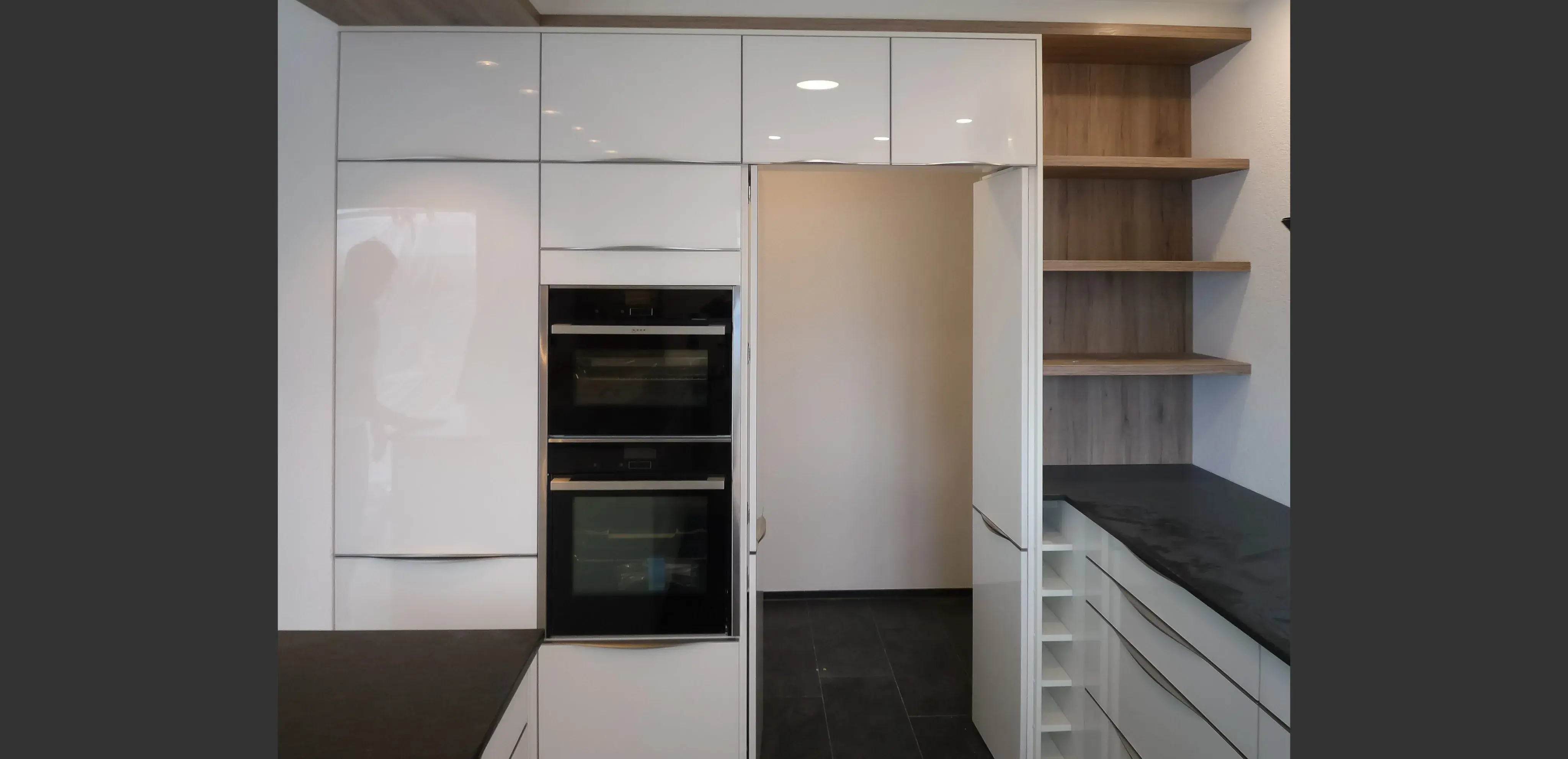 Moderne weiße Küche mit Einbauöfen, offenen Regalen und dunkler Arbeitsplatte.