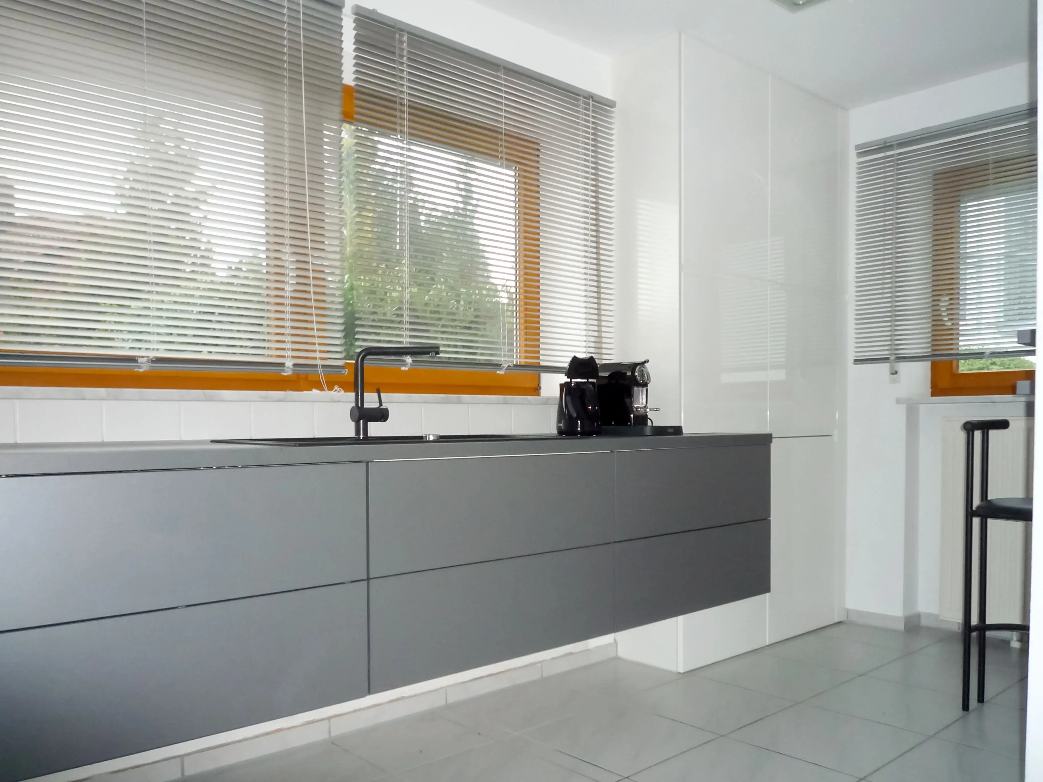 Moderne Küche mit grauen Schränken, großen Fenstern und Kaffeemaschine.