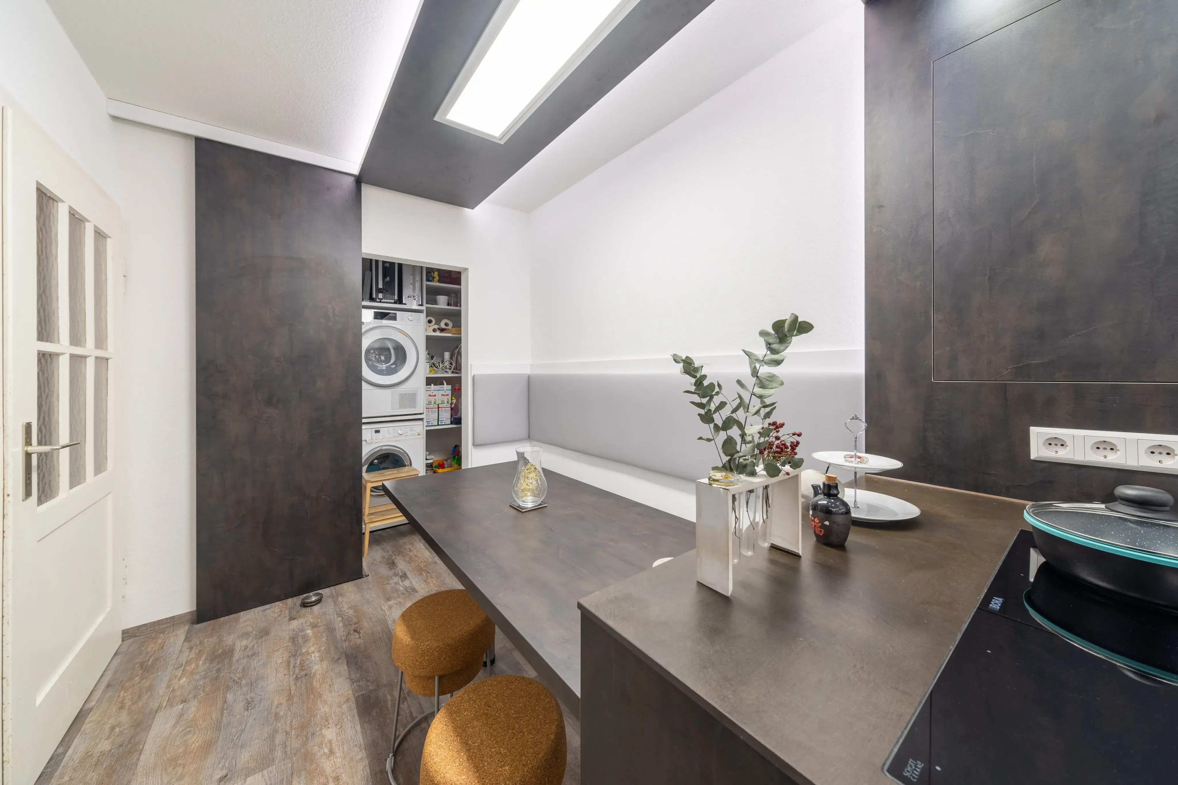 Moderne Küche mit dunklen Schränken, Waschtrockner, langer Tisch und drei Hockern.