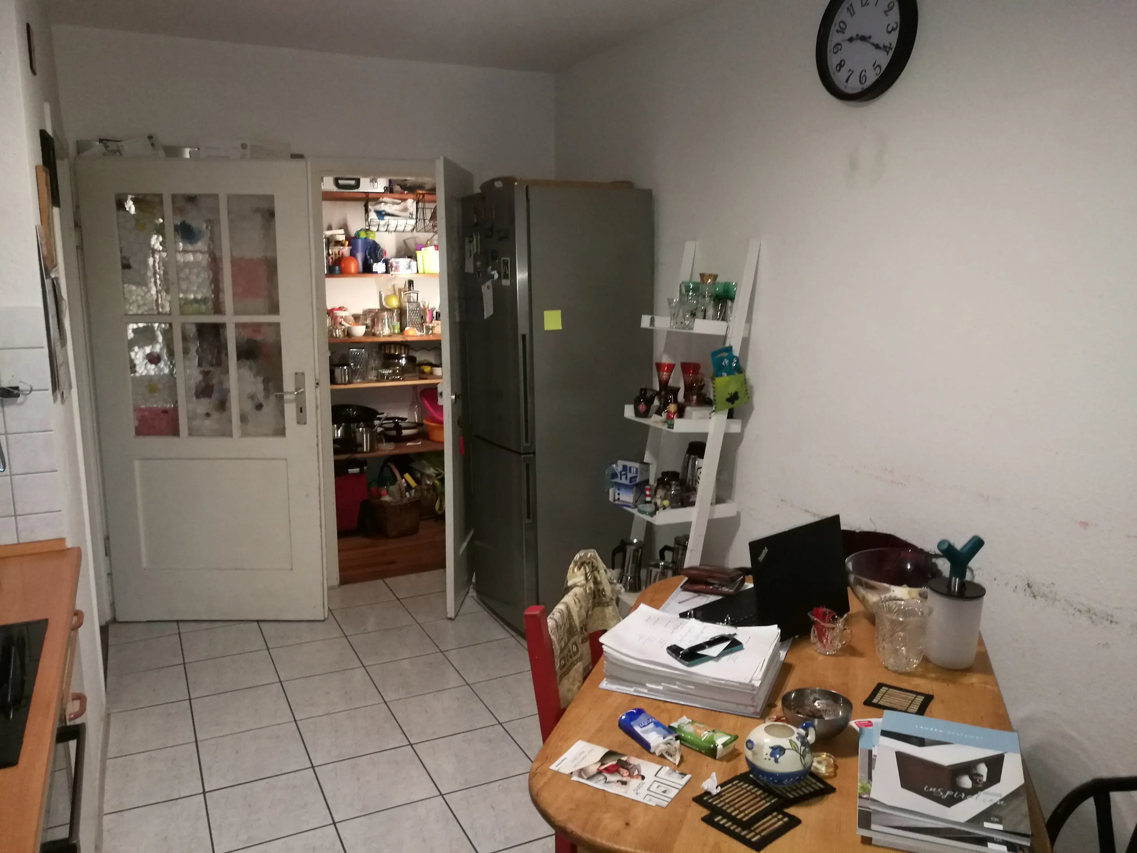 Küche mit Esstisch, viel Papierkram, Kühlschrank und offener Speisekammer.