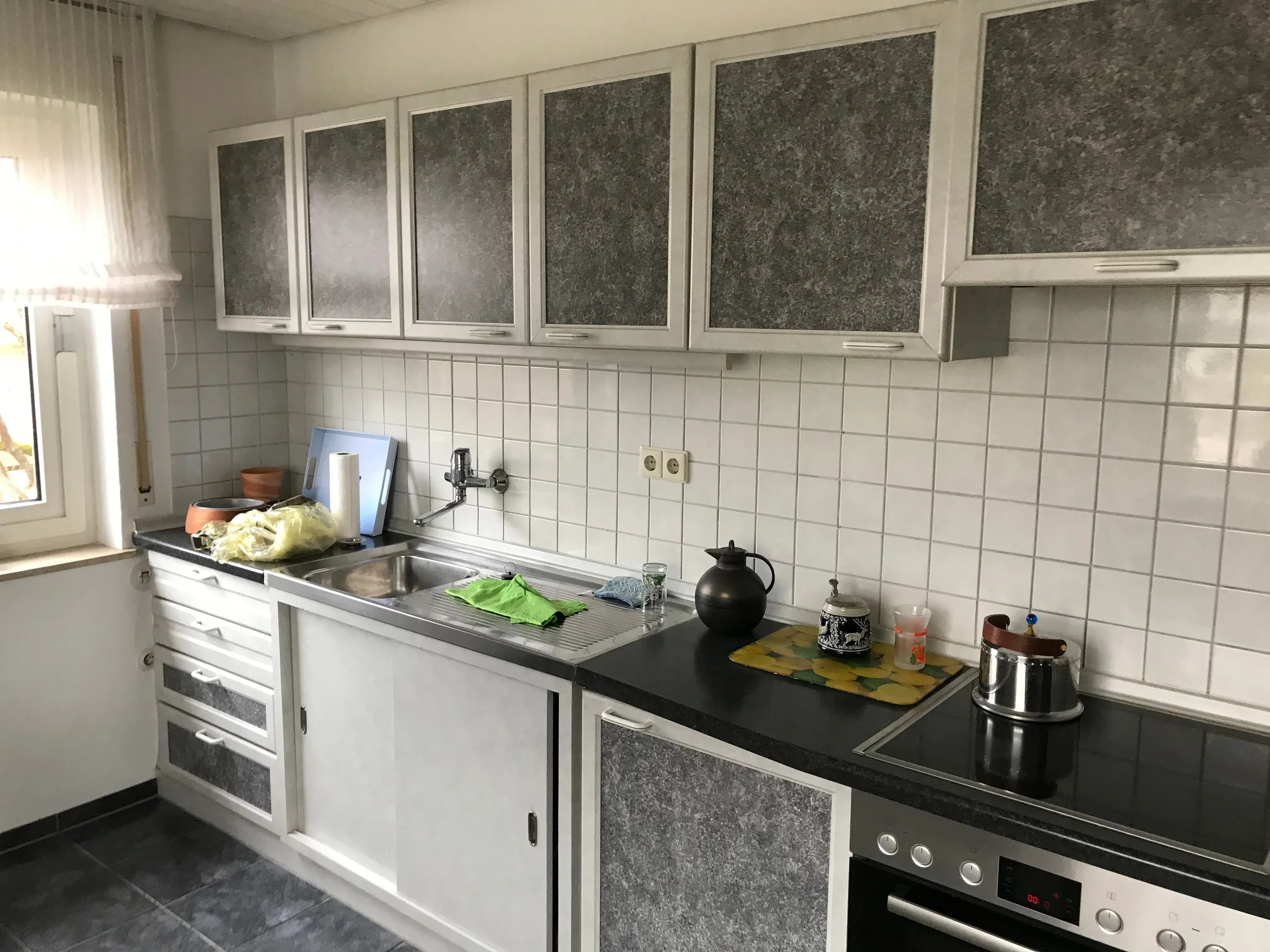 Helle Küche mit grauen Schränken, weißem Fliesenspiegel und Haushaltsgegenständen auf der Arbeitsplatte.